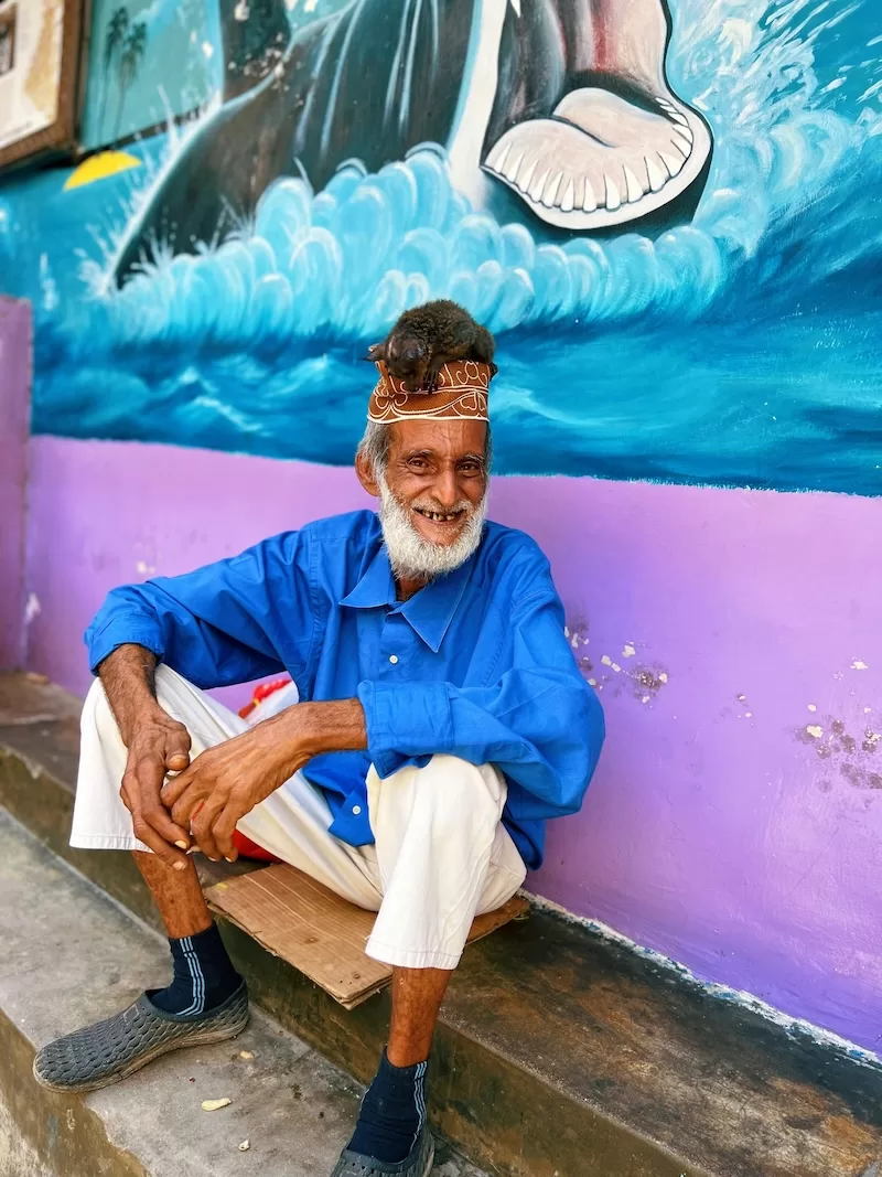 A man in a blue shirt and traditional cap sits smiling in front of a colorful mural of a shark at Jaw’s Corner, a famous social hub in Stone Town, Zanzibar.