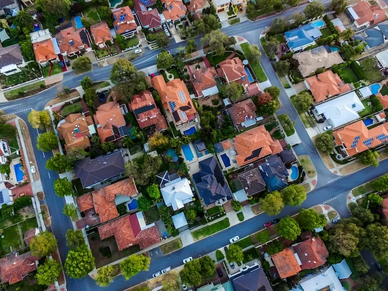High-angle aerial view of a suburban neighborhood with red-roofed houses and backyard swimming pools, symbolizing the traditional but costly aging-in-place model.