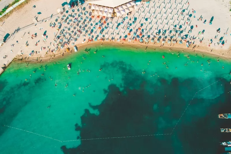 High-angle aerial view of a crowded beach with vibrant turquoise water and many colorful umbrellas, representing a popular coastal destination like Bulgaria.