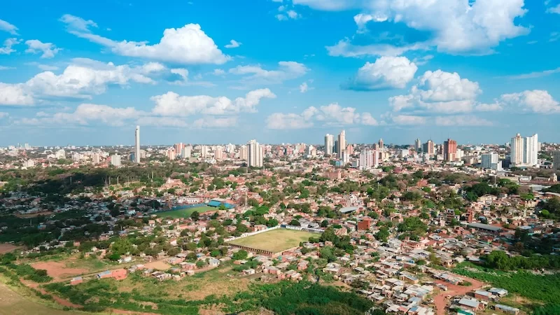 A wide aerial view of a sprawling city with a mix of low-rise residential buildings and modern skyscrapers under a bright blue sky with fluffy white clouds.