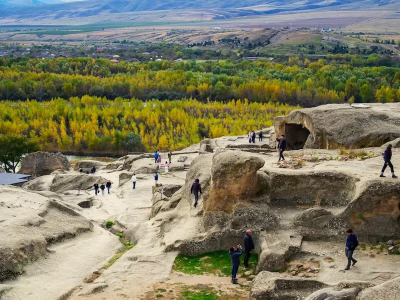 People explore an ancient cave city carved into rocky cliffs, with a lush valley of yellow and green trees in the background, representing Georgia's landscape.