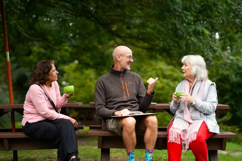 Three older adults sit at a wooden picnic table in a lush green park, smiling and talking over coffee, illustrating the social integration of expat life.