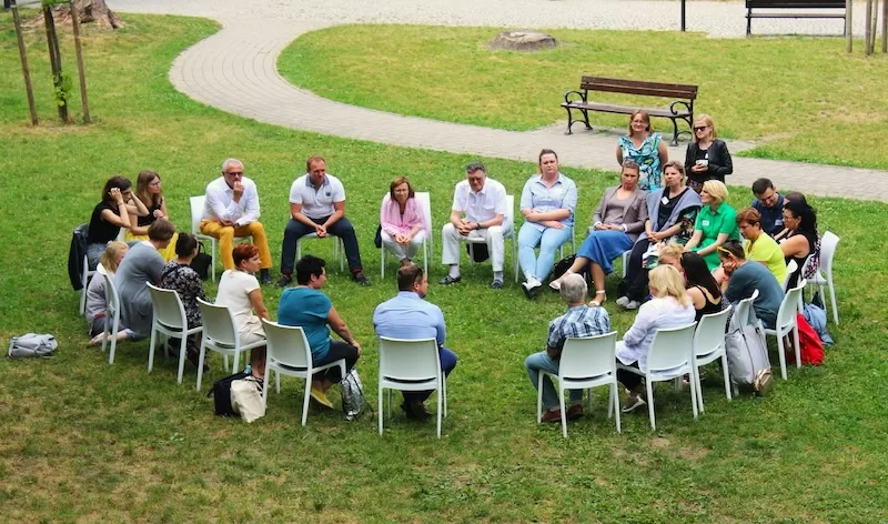 A large group of people sits in a circle of white chairs on a green lawn for an outdoor community meeting, representing the strong social fabric of expat hubs.
