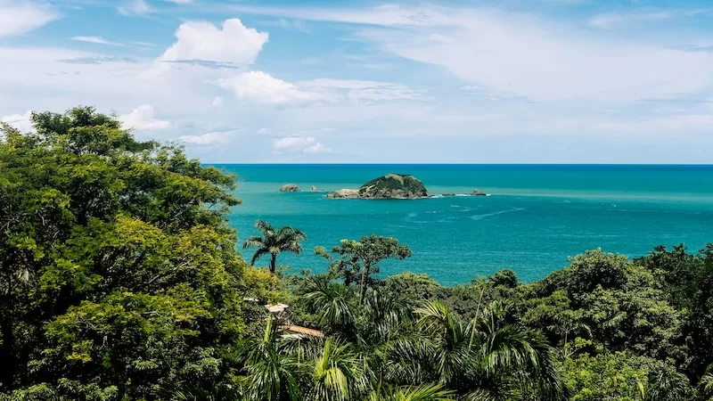 A breathtaking view of a small island in the middle of a turquoise ocean, seen through lush green tropical trees, representing the Pura Vida lifestyle in Costa Rica.