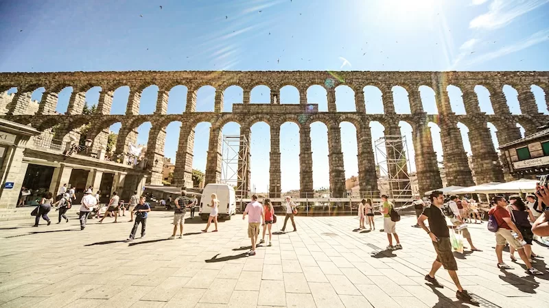A wide-angle view of the ancient Roman Aqueduct of Segovia in Spain, with people walking in the sun-drenched plaza under a clear sky with birds flying.