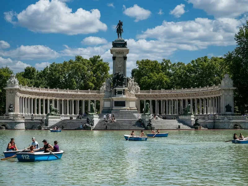 A wide view of the Monument to Alfonso XII in Retiro Park, Madrid, with people rowing blue boats on the calm lake in front of the grand colonnade.