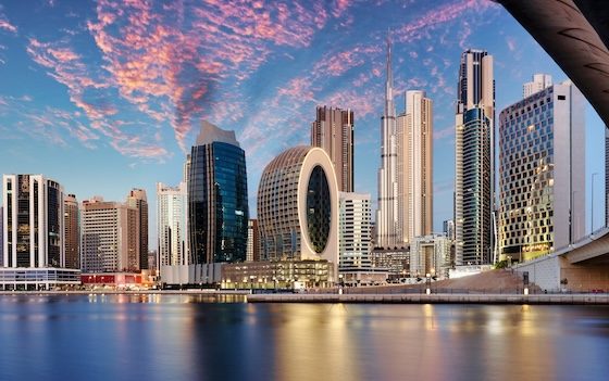 Iranian flag waving on a pole with Tehran cityscape and mountains in background, representing Iran-US geopolitical tensions and Middle East conflict