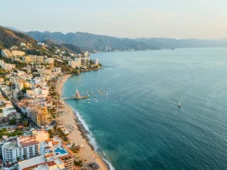 Aerial view of Puerto Vallarta coastal town with turquoise ocean water, sandy beaches, white buildings, green mountains, and boats anchored in the bay