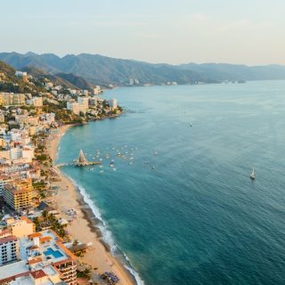 Aerial view of Puerto Vallarta coastal town with turquoise ocean water, sandy beaches, white buildings, green mountains, and boats anchored in the bay