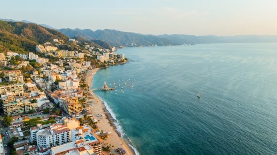 Aerial view of Puerto Vallarta coastal town with turquoise ocean water, sandy beaches, white buildings, green mountains, and boats anchored in the bay