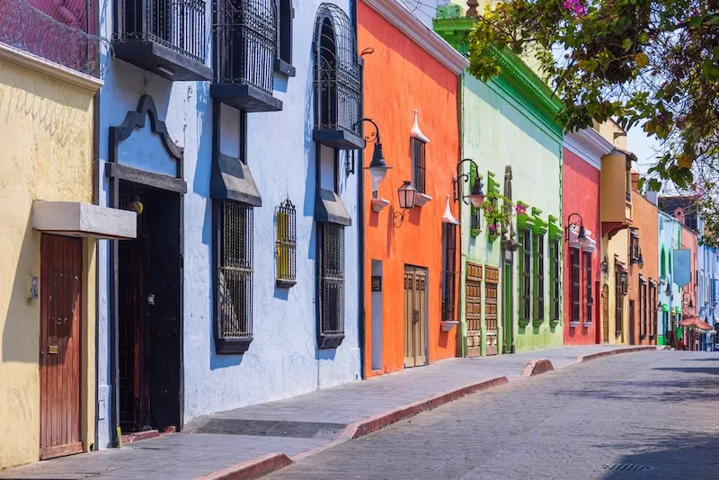 A vibrant Mexican street lined with colorful colonial buildings in shades of blue, orange, green, and red, featuring traditional wrought iron window grilles