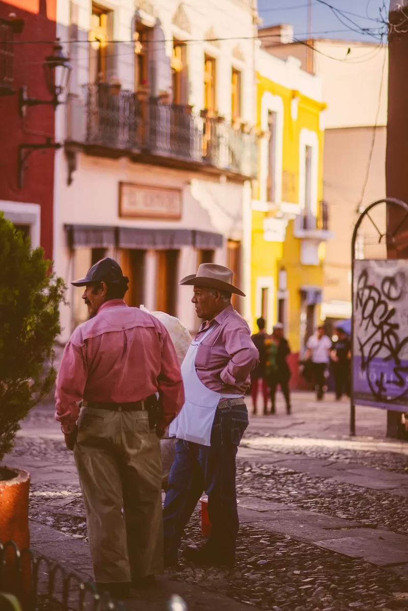 Two men in cowboy hats and pink shirts stand on a colorful Mexican colonial street with red, yellow, and cream buildings with traditional architecture
