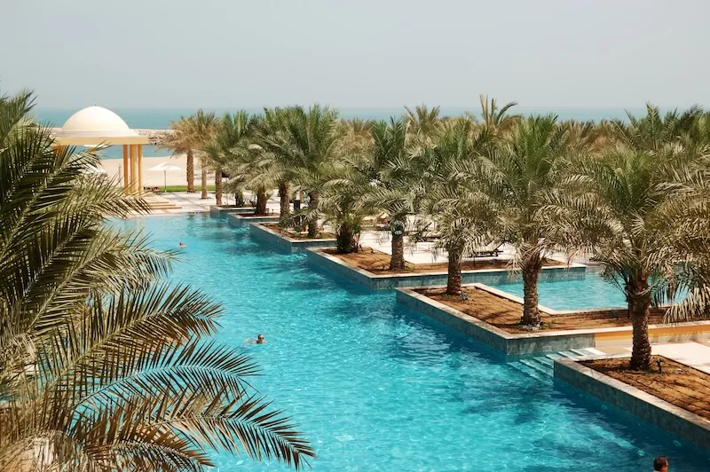 Luxury beachfront resort with palm trees and turquoise swimming pools overlooking the Arabian Gulf.