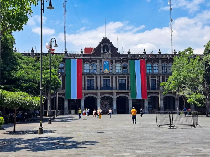 A historic Mexican government building with large national flags draped from the facade, overlooking a paved public square with people walking on a sunny day