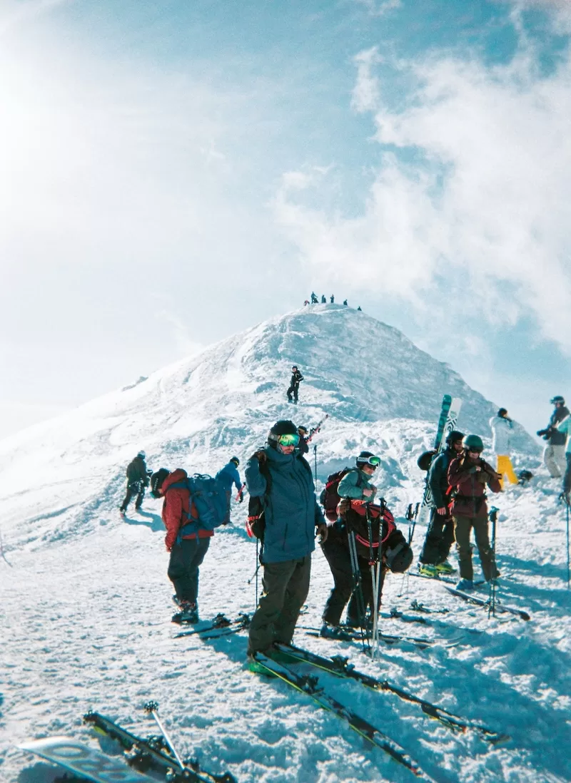 Skiers and mountaineers standing on a snowy mountain ridge under a bright winter sky.
