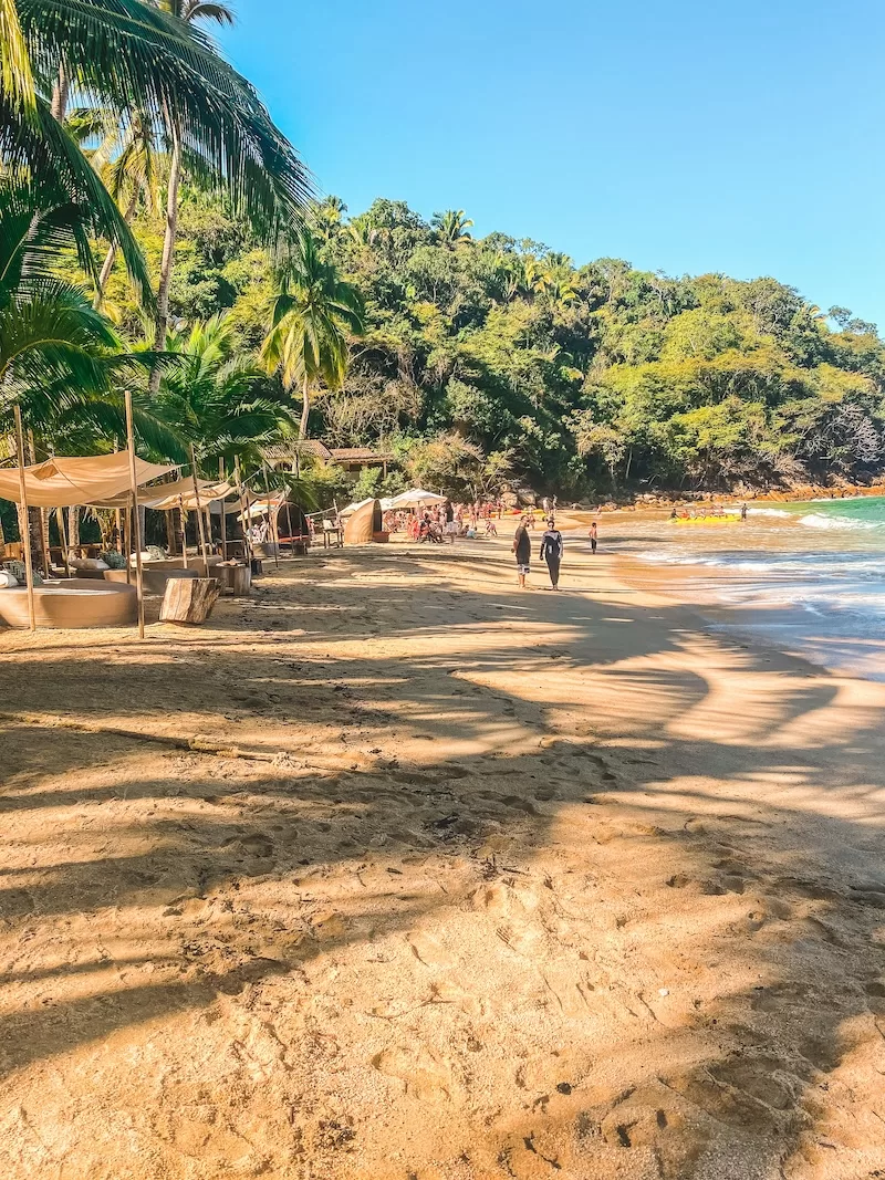 Scenic beach with golden sand, palm tree shade structures, turquoise ocean waves, green jungle forest, and people enjoying the tropical seaside view