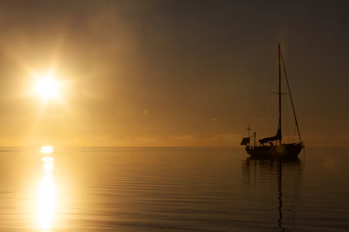 Sailboat anchored on calm ocean water at sunset with golden sunlight reflecting across the sea.