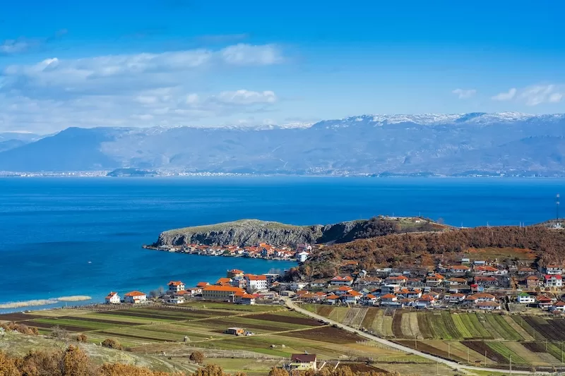 Rural village and farmland beside a bright blue lake in North Macedonia with mountains in the distance.