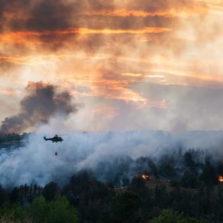 Helicopter carrying water bucket over a smoky forest wildfire at sunset.