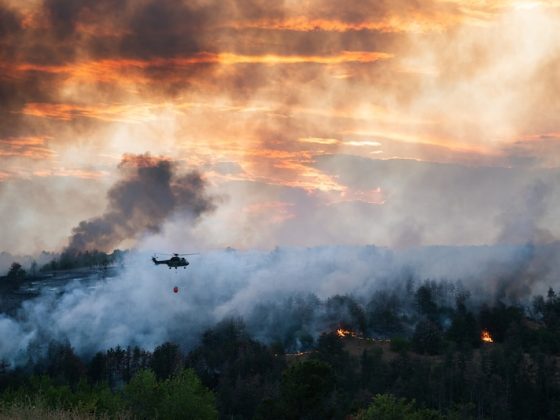 Helicopter carrying water bucket over a smoky forest wildfire at sunset.