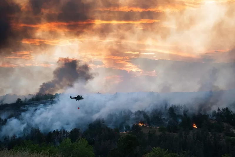 Helicopter carrying water bucket over a smoky forest wildfire at sunset.