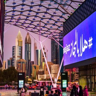 Evening street scene in Dubai with illuminated digital billboards, pedestrians, and modern skyscrapers rising in the background