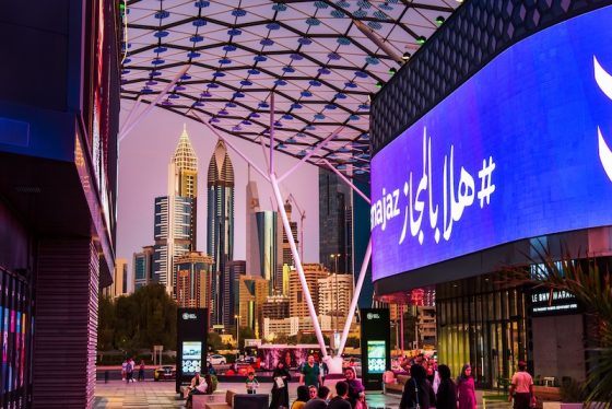 Evening street scene in Dubai with illuminated digital billboards, pedestrians, and modern skyscrapers rising in the background