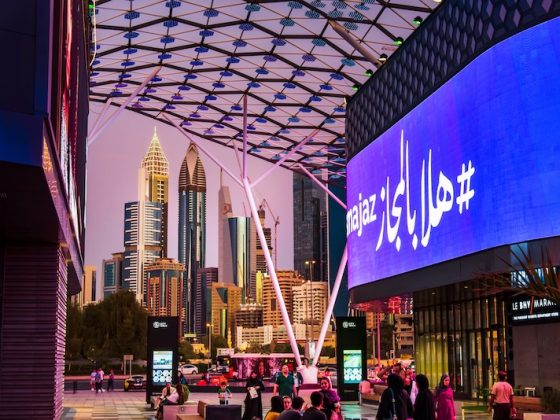 Evening street scene in Dubai with illuminated digital billboards, pedestrians, and modern skyscrapers rising in the background