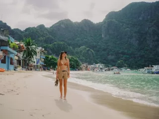 Woman walking barefoot along a beach in the Philippines with boats, waterfront buildings, and lush limestone hills in the background