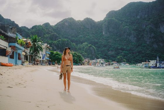 Woman walking barefoot along a beach in the Philippines with boats, waterfront buildings, and lush limestone hills in the background