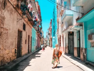 Woman walking down a narrow street in Havana lined with weathered pastel buildings beneath a bright blue sky