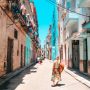 Woman walking down a narrow street in Havana lined with weathered pastel buildings beneath a bright blue sky