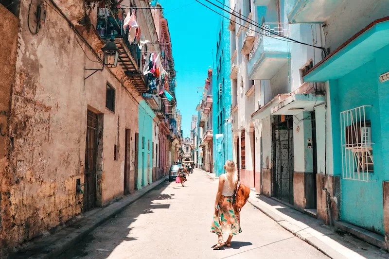 Woman walking down a narrow street in Havana lined with weathered pastel buildings beneath a bright blue sky