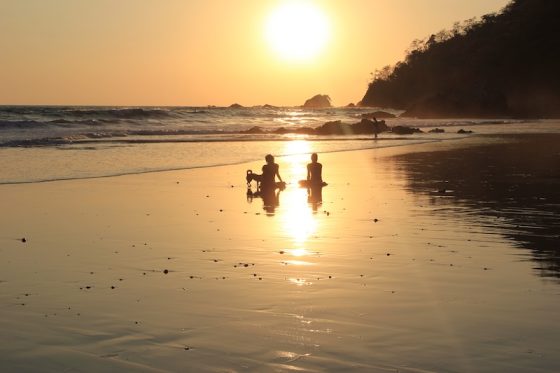 Two people sitting on a beach at sunset with waves rolling in and rocky coastline silhouetted against the golden sky