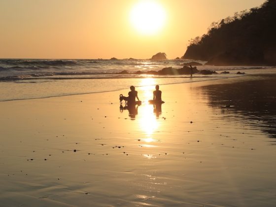 Two people sitting on a beach at sunset with waves rolling in and rocky coastline silhouetted against the golden sky