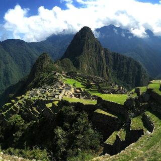 Wide view of Machu Picchu with green terraced ruins set high among dramatic Andes mountains under a bright sky