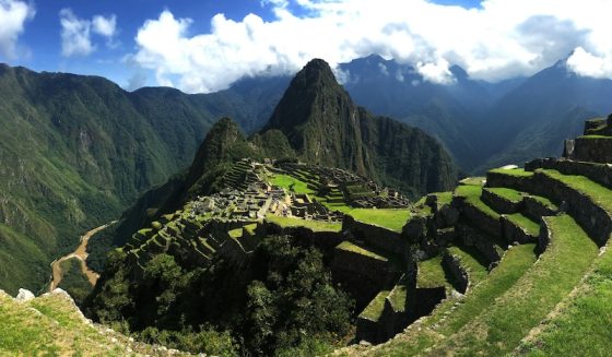 Wide view of Machu Picchu with green terraced ruins set high among dramatic Andes mountains under a bright sky