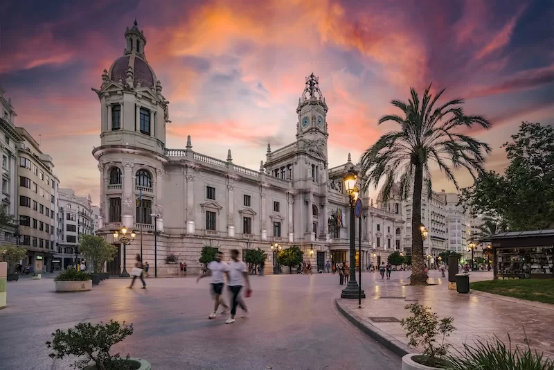 Historic civic building and palm-lined square in Valencia at sunset with people walking through the plaza