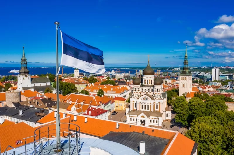 Estonian flag flying above Tallinn’s old town skyline with historic churches and orange rooftops below.