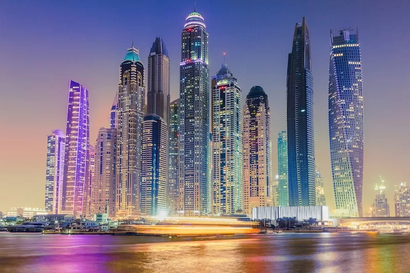 Night view of Dubai Marina skyline with illuminated skyscrapers reflected on the water and a blurred boat passing in the foreground