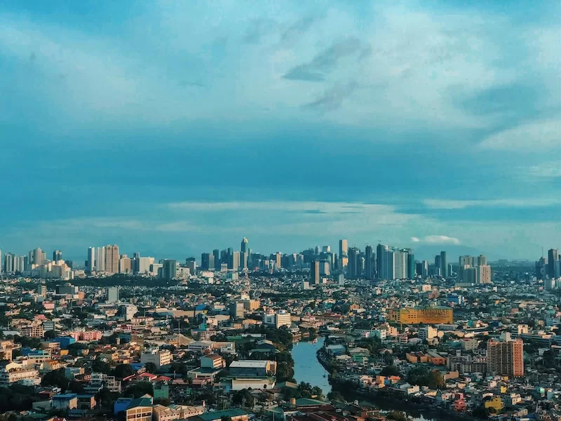 Wide view of the Manila skyline with dense urban neighborhoods, river channels, and modern high-rise buildings under a cloudy sky