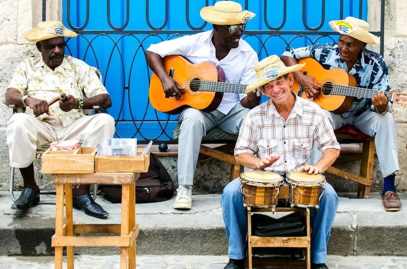 Group of Cuban street musicians playing guitars and hand drums while seated on a sidewalk in front of a bright blue doorway