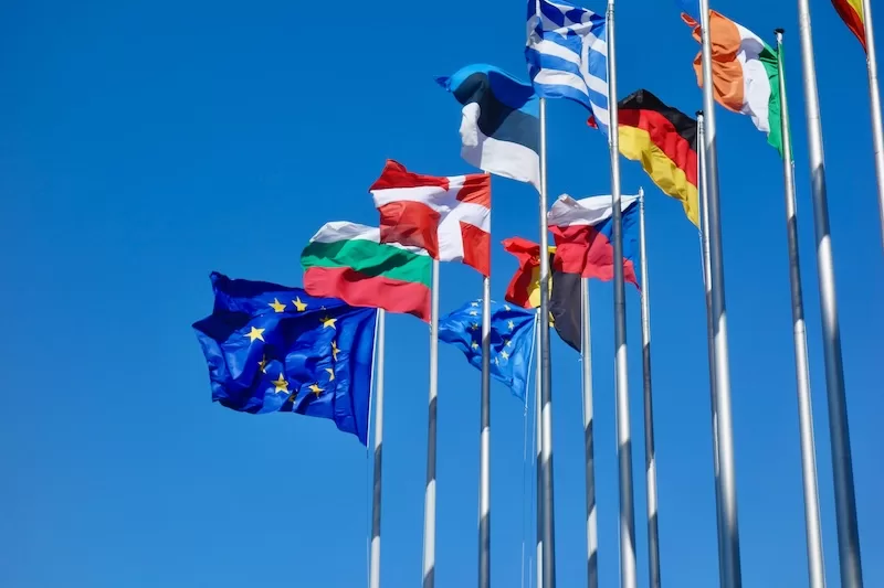 European Union and national flags flying together on tall flagpoles against a clear blue sky.