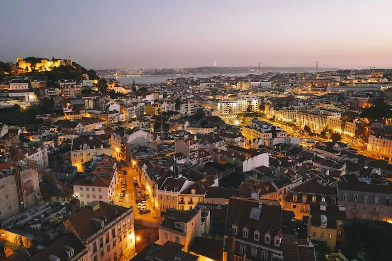Elevated dusk view over Lisbon with terracotta rooftops, glowing streetlights, historic buildings, and the river in the background