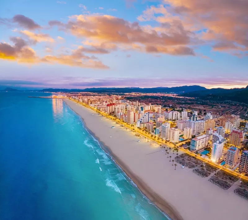Aerial view of Valencia’s coastline at dusk with sandy beach, turquoise sea, lit promenade, and city buildings stretching along the shore