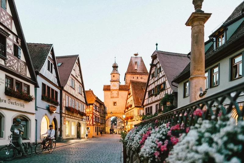Historic German old town street lined with half-timbered buildings, bicycles, flowers, and a clock tower archway at dusk