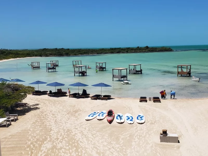 Sandy beach in Belize with paddleboards, lounge chairs, blue umbrellas, and overwater cabanas set in calm shallow turquoise water