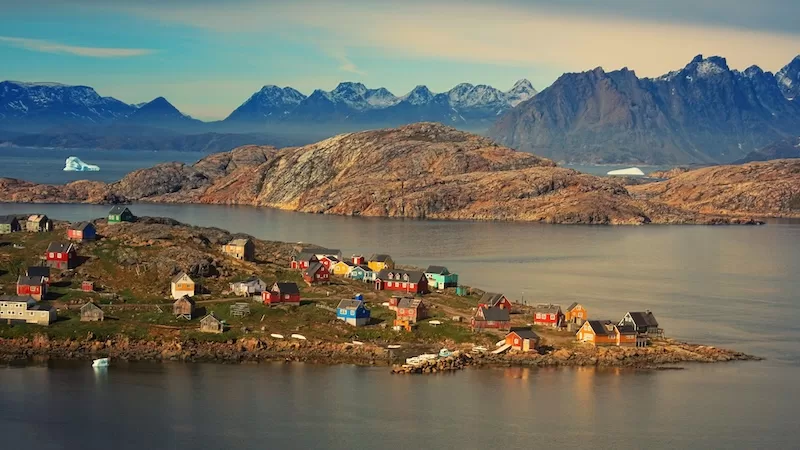 Small colorful settlement on a rocky Arctic shoreline with calm water, snow-dusted mountains, and icebergs in the distance