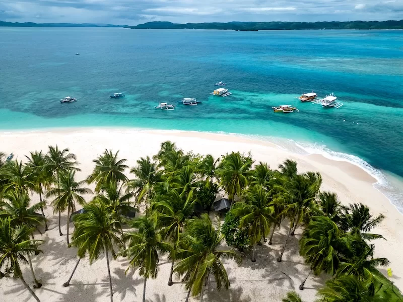 Aerial view of a white-sand beach in the Philippines lined with palm trees and small boats floating in bright turquoise water