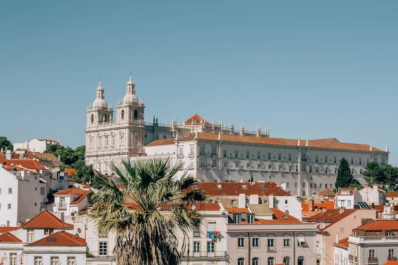 View over Lisbon rooftops toward a large white monastery building beneath a clear blue sky